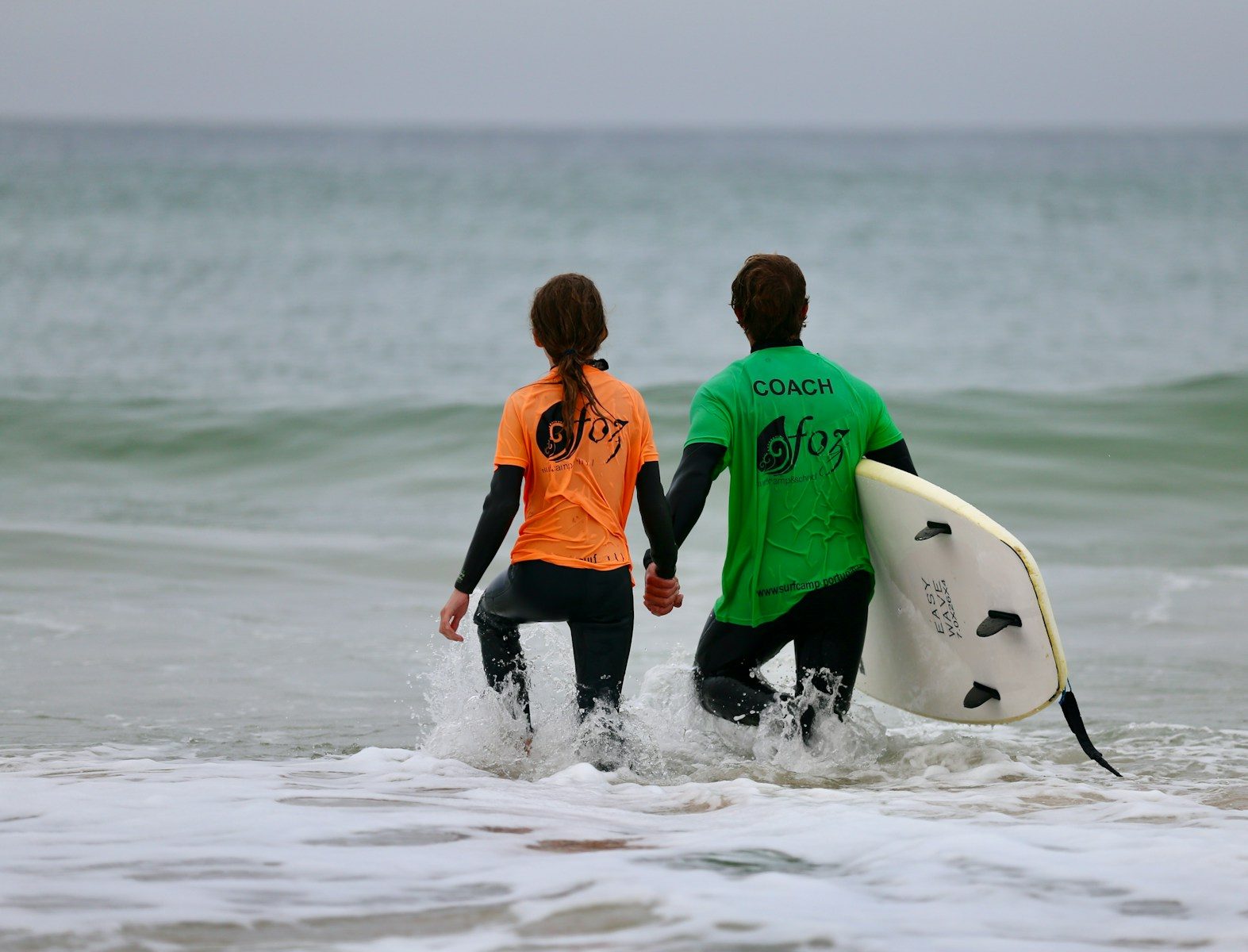 Two surfers holding hands walk into the ocean.